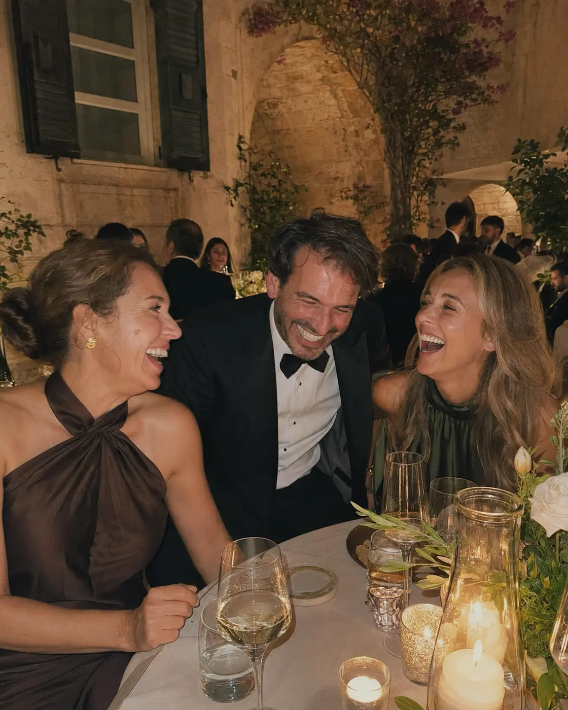 Three friends laughing over dinner in a stone courtyard
